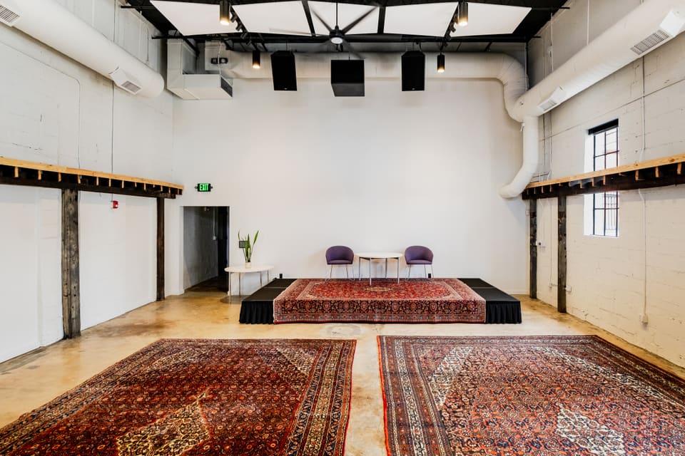 The same multi-purpose space after renovation, with white walls, a polished wood floor, an exposed black-steel truss ceiling, and layered area rugs.